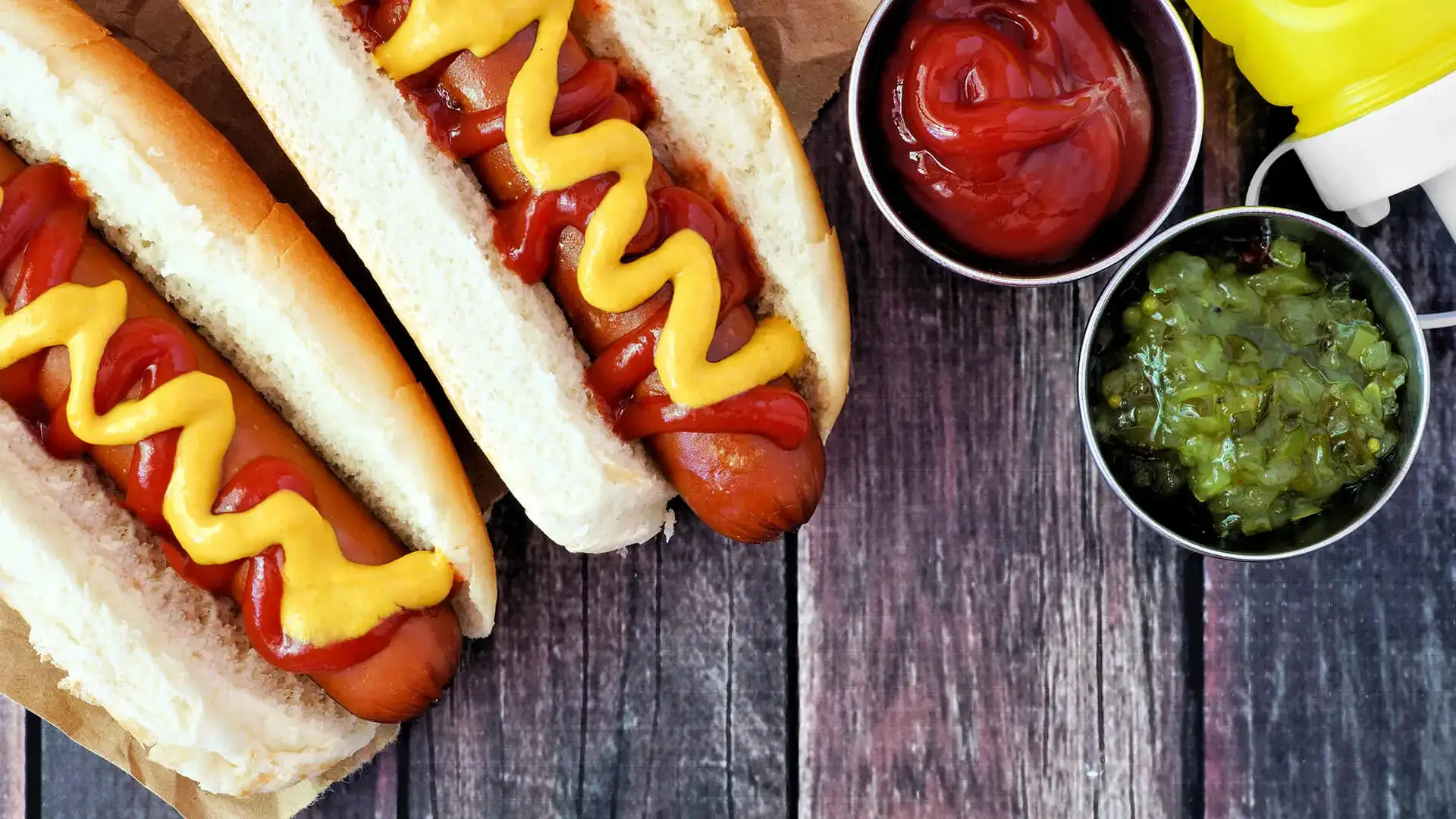 Image of two hot dogs with sides of ketchup and relish.