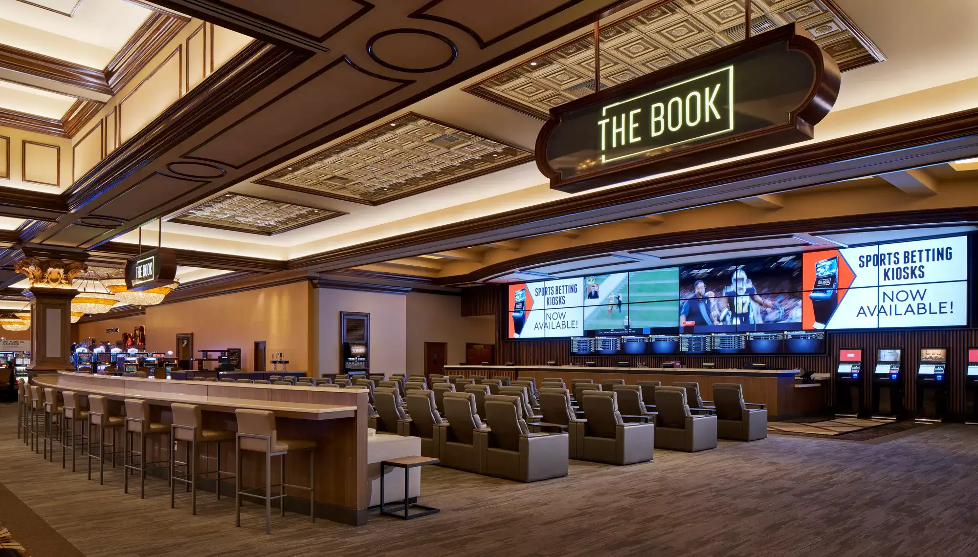 Interior view of The Book sportsbook with stadium-style seating, large video screens displaying sporting events, and self-service betting kiosks