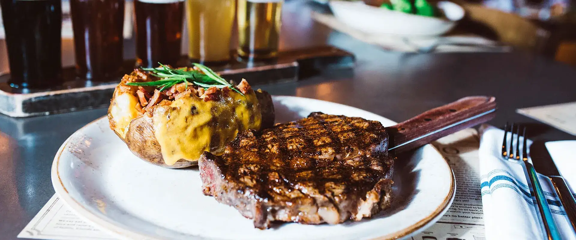 Steak and baked potato on a plate with flight of beer in the background.