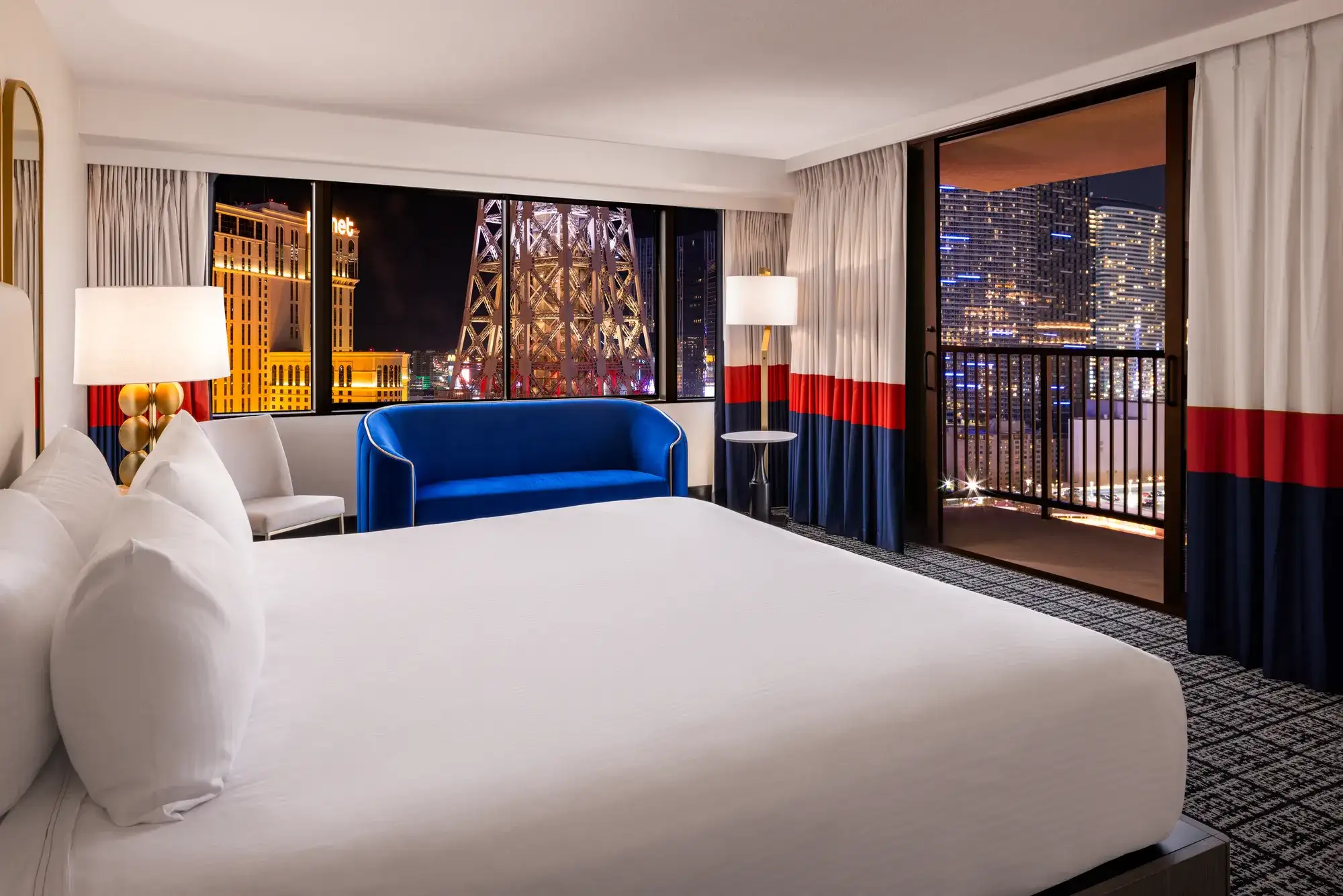 Paris Las Vegas Balcony Room at night with a king bed, blue velvet sofa, private balcony, and Center Strip view of the Eiffel Tower replica - the kind of Vegas hotel room travelers do not regret booking.