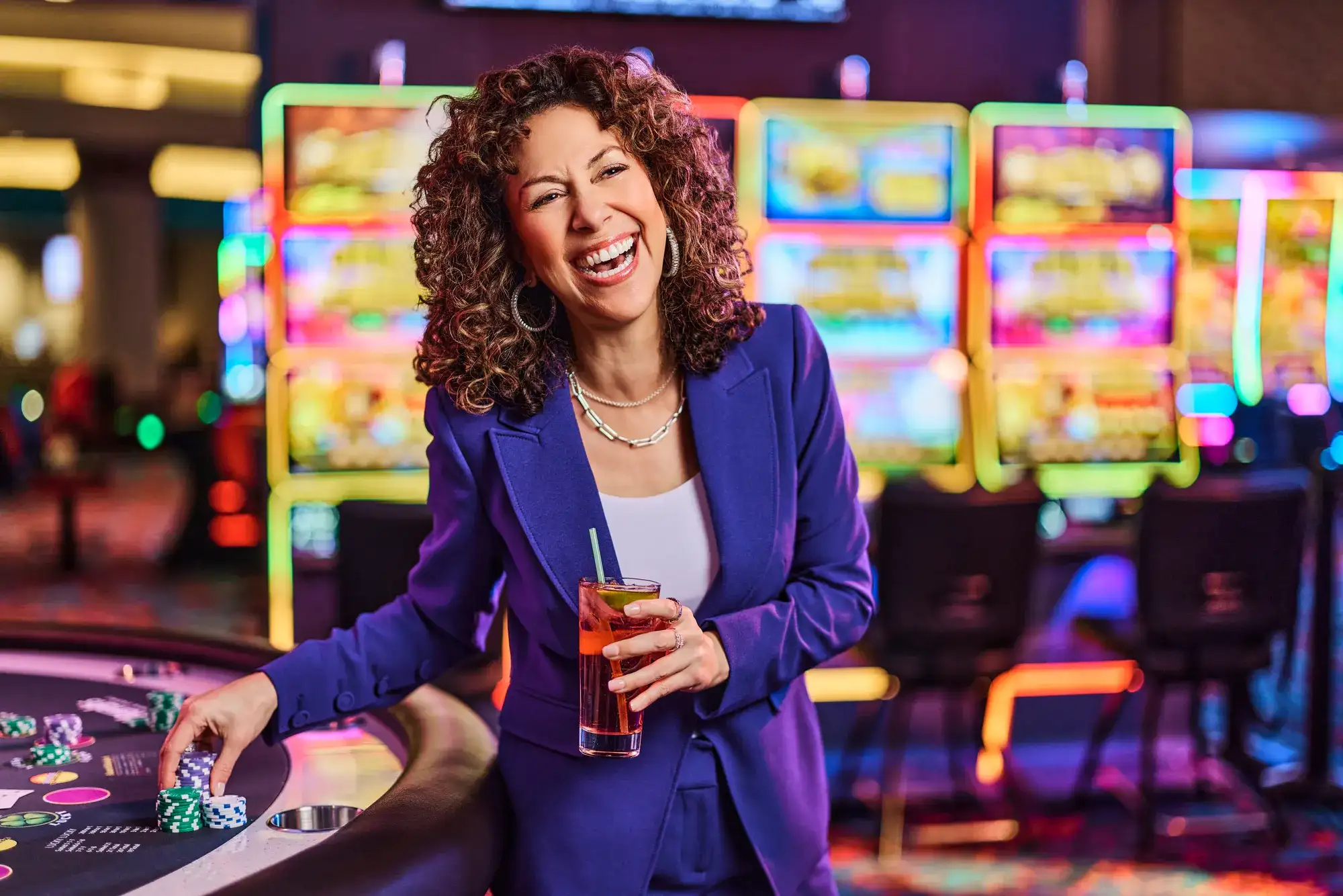 A casino guest laughing and holding a cocktail at a Las Vegas casino table game with colorful neon slot machine displays glowing in the background