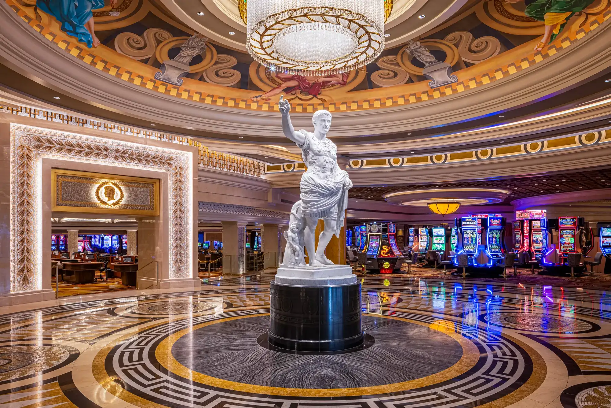 Caesars Palace casino interior with a classical statue, crystal chandelier, marble floors, gaming tables, and illuminated slot machines