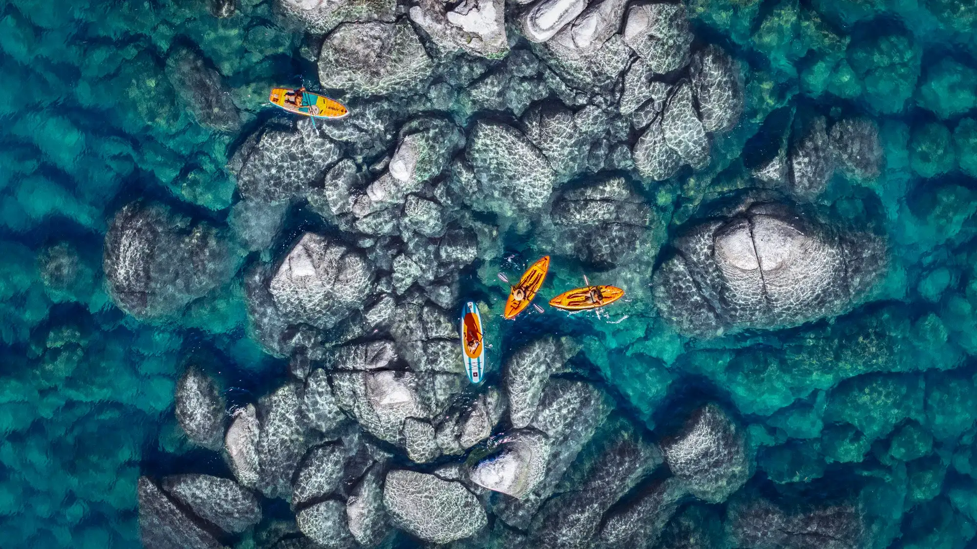 Aerial view of paddleboarders and kayakers gliding over Lake Tahoe's crystal-clear turquoise water in summer, with submerged granite boulders visible beneath the surface near the shoreline.