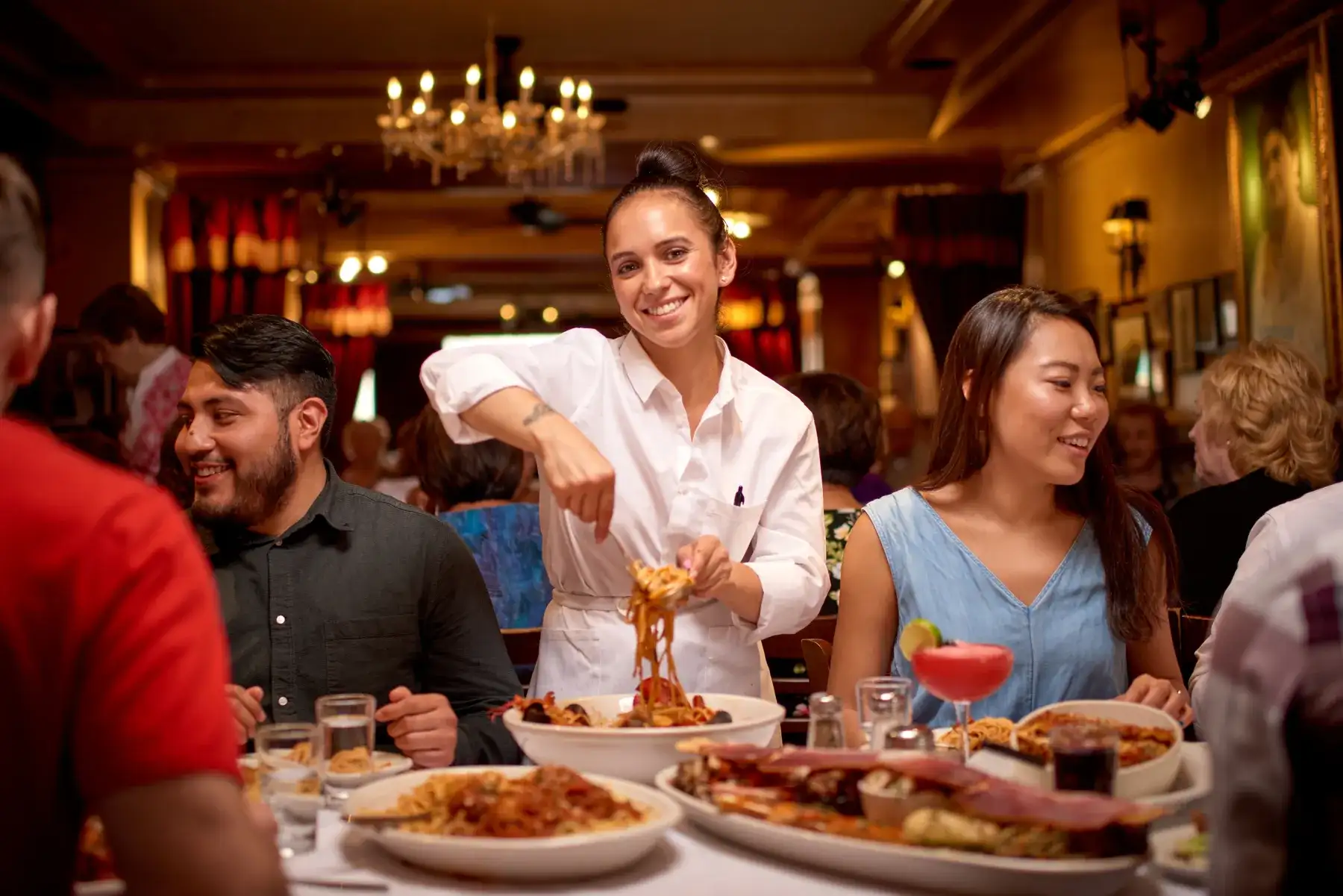 Server mixing pasta in a large serving bowl at a restaurant table with guests, shared dishes, and a cocktail