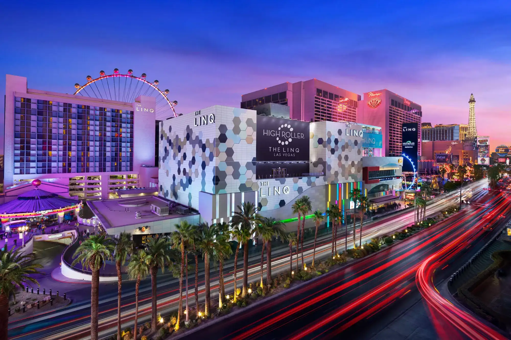 Night view of Center Strip with illuminated resort buildings, an observation wheel, palm trees, and traffic light trails.