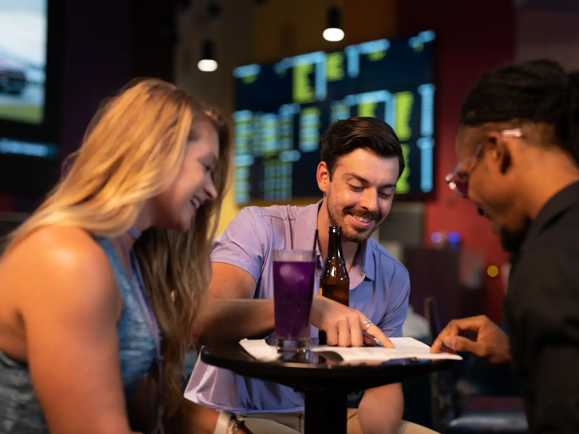 4 guests review a paper at a sportsbook table beneath betting screens
