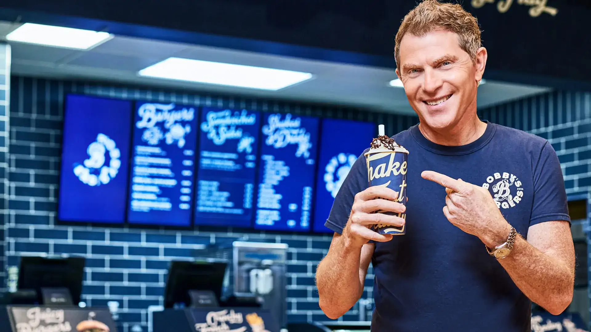 Chef Bobby Flay holding a shake in front of Bobby's Burgers.