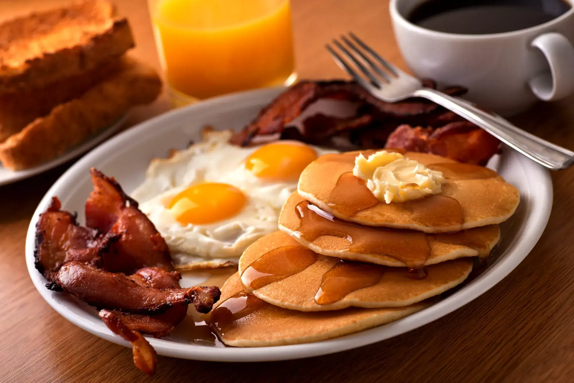 A plate of pancakes, eggs and bacon with orange juice, coffee and toast in the background