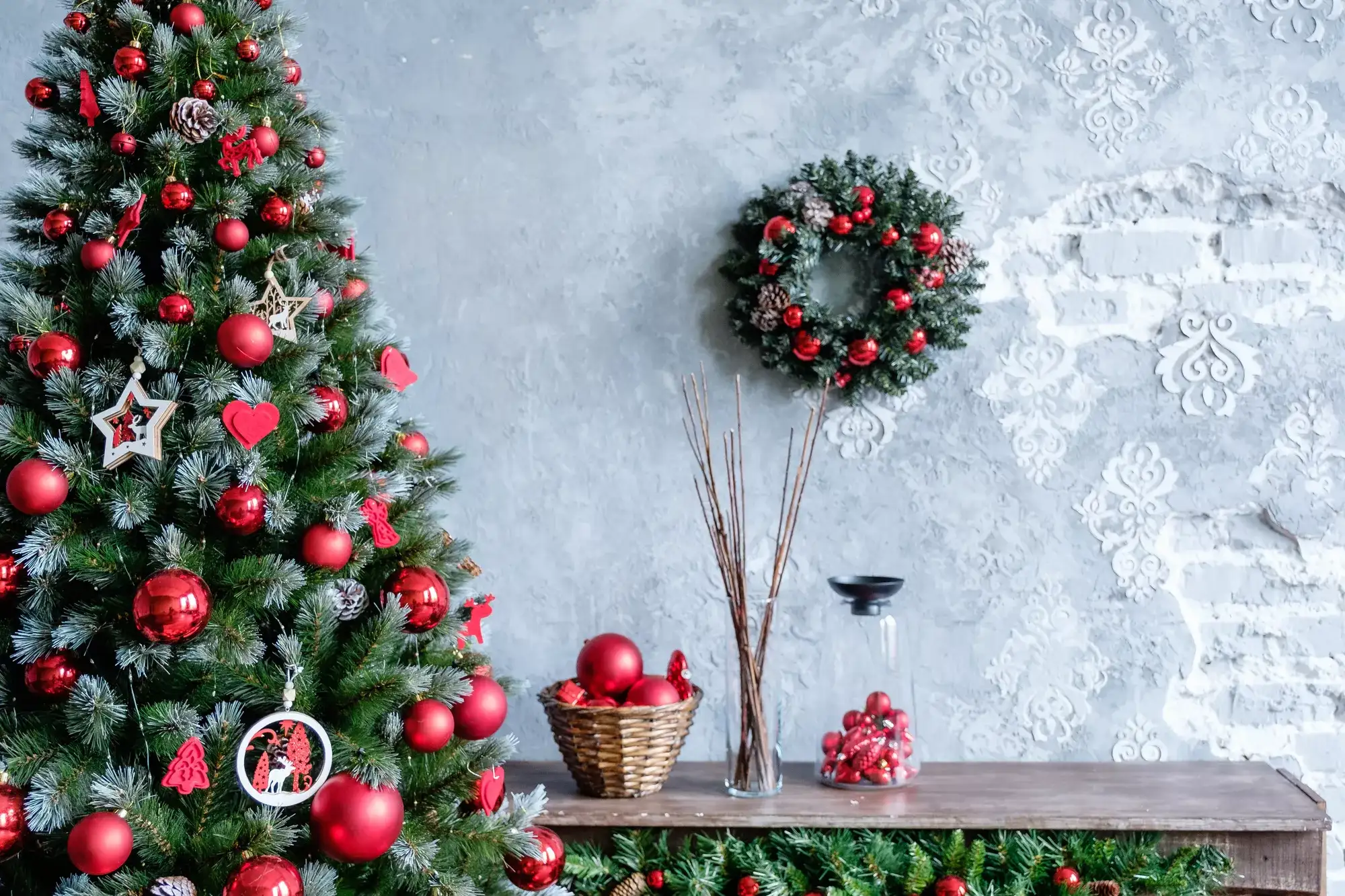 Christmas tree and matching wreath with red ornaments beside a holiday-decorated wooden table