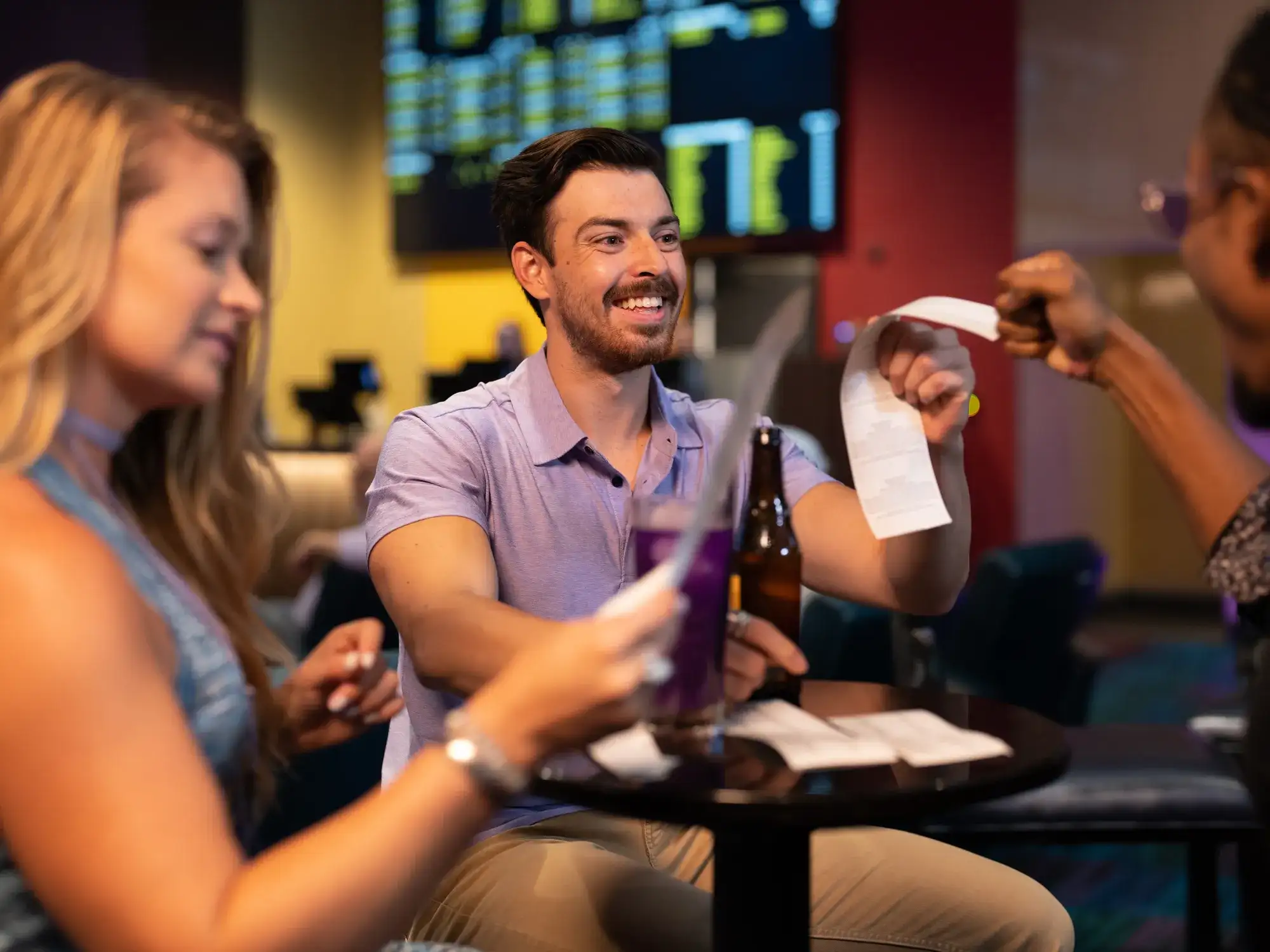 3 guests show off a paper at a sportsbook table beneath betting screens
