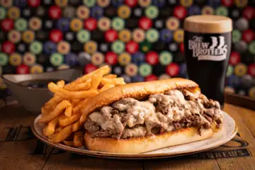 Philly cheesesteak and fries on a plate with a glass of dark beer on a table at The Brew Brothers.