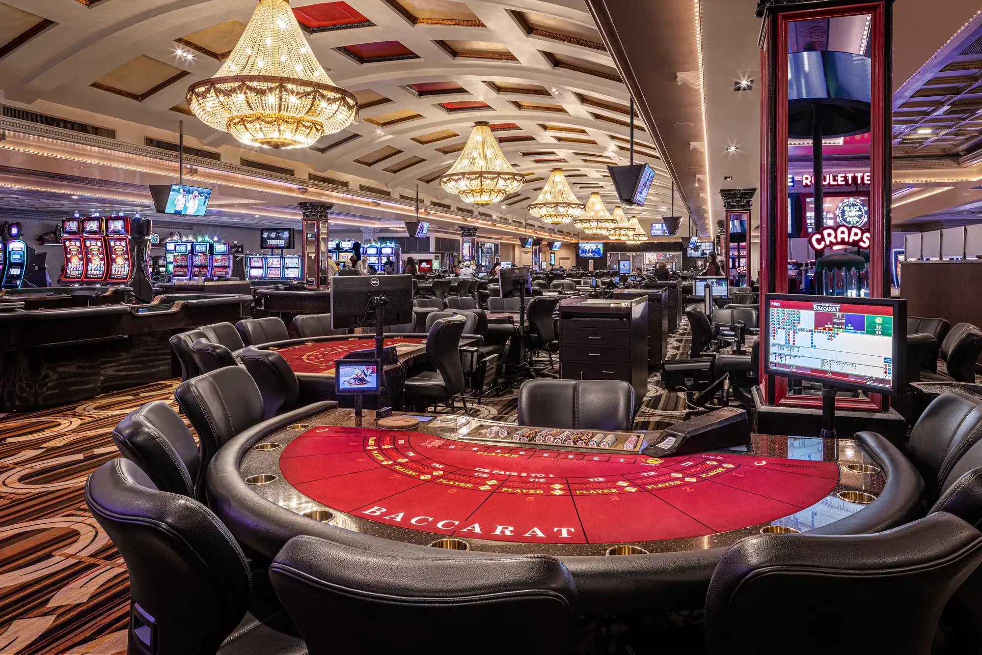 Red baccarat tables surrounded by black chairs on a casino floor with slot machines, gaming tables, and illuminated chandeliers